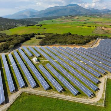 Aerial view of a solar farm in a lush rural area, featuring rows of photovoltaic panels oriented toward the sun with mountains in the background