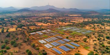 Aerial view of a rural landscape with solar panels and a wind turbine, showcasing sustainable energy in a developing region. Houses and farms dot the dry, arid landscape.